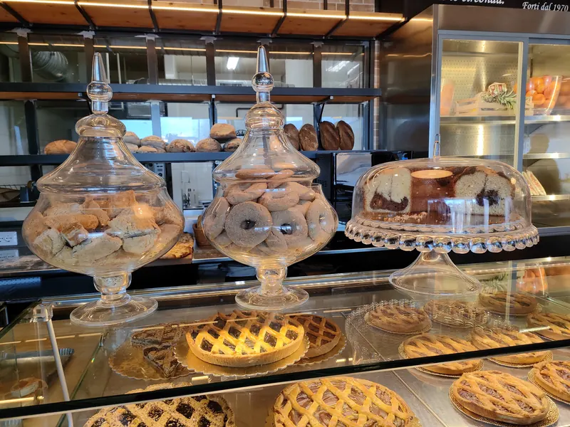 Il banco di Forti: ciambelle e biscotti nei vasi di vetro, una torta marmorizzata sotto la campana, crostate in vetrina e pagnotte sugli scaffali